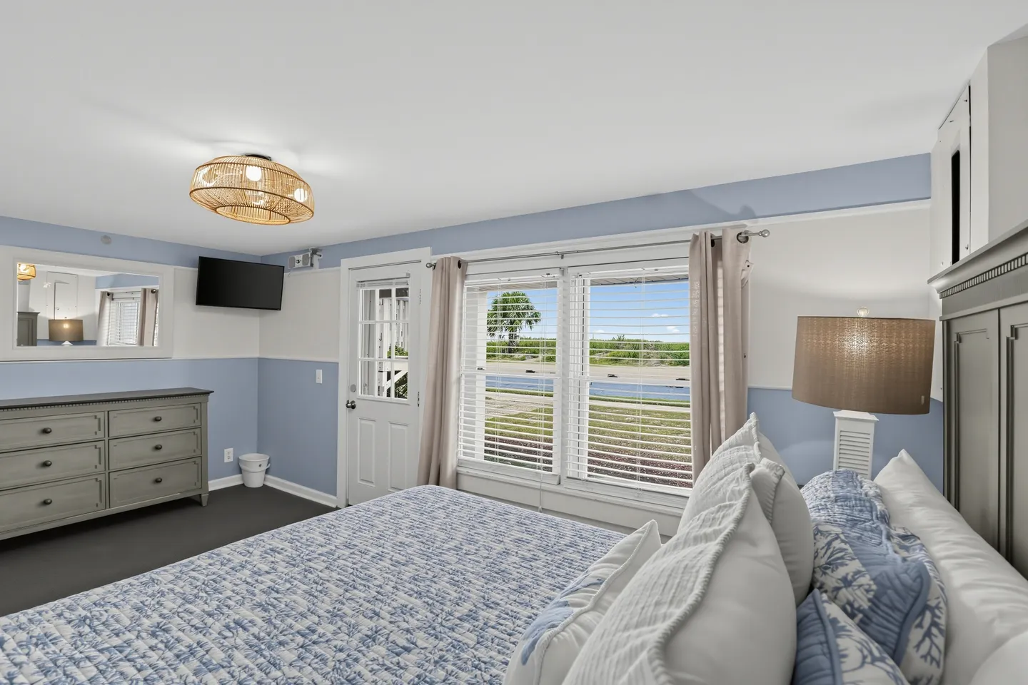 Bedroom with blue and white walls, a made bed, a wooden dresser, and large windows looking outside.