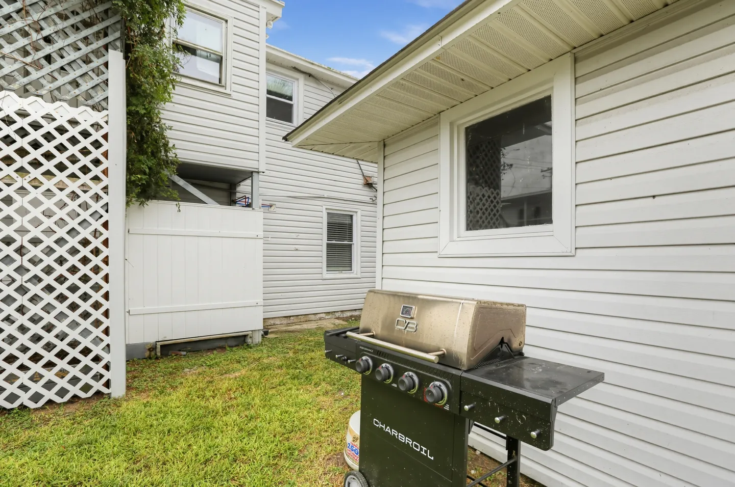 A Char-Broil freestanding barbecue grill set outside on a patch of grass between white vinyl-sided exterior walls.