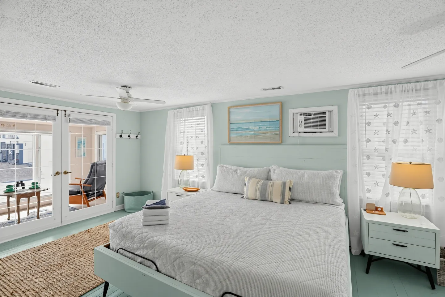 Bedroom with light aqua walls, a large bed with a white quilted spread, matching nightstands with lamps, and French doors opening to an enclosed porch.
