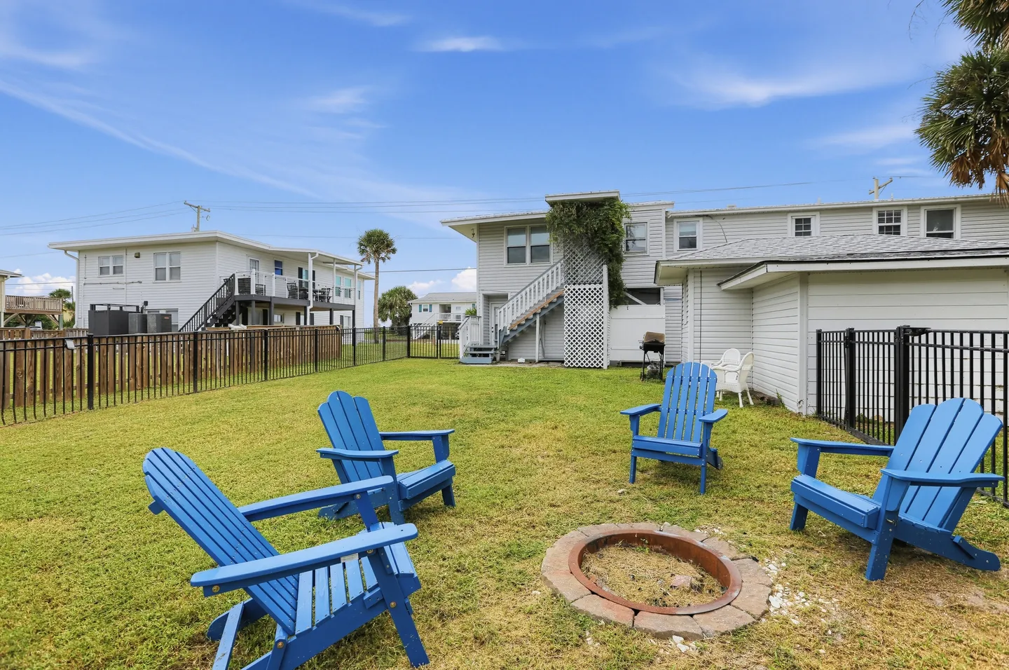 A grassy backyard with a sunken metal-lined fire pit encircled by four bright blue Adirondack chairs, enclosed by black and wooden fencing with nearby buildings under a clear blue sky.