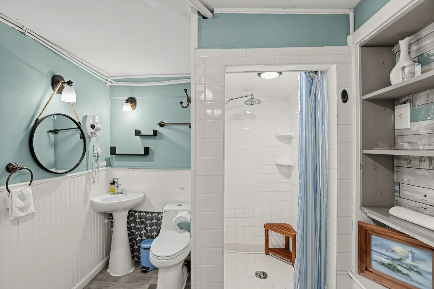 Bathroom with light blue walls, a white pedestal sink, toilet, round hanging mirror, open shelving, and a tiled walk-in shower.
