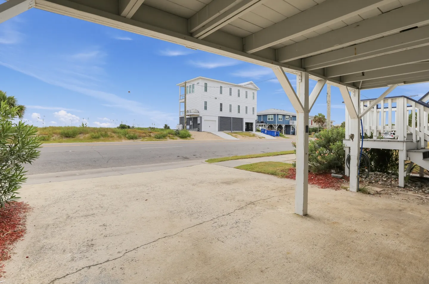 Covered concrete carport underneath a home looking out toward a paved road, sandy mounds, and neighboring coastal houses under a blue sky.