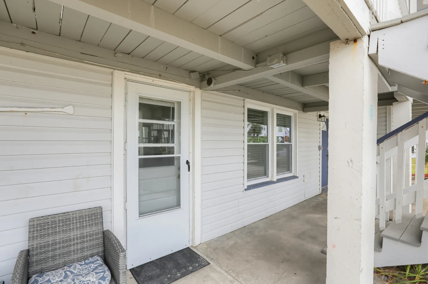 A covered concrete entryway featuring white siding, a white front door with a window, windows, and an outdoor chair.