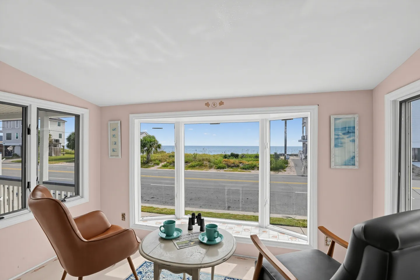 Seating area with two armchairs and a small table in front of a bay window overlooking a road and the ocean.