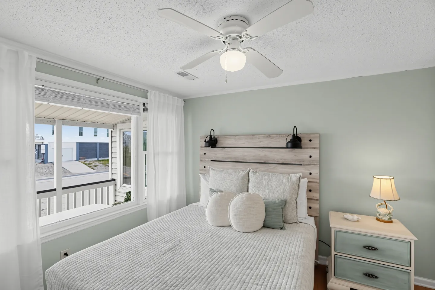 Bedroom with a made bed, rustic wooden headboard, bedside table with lamp, and windows looking out to a porch.