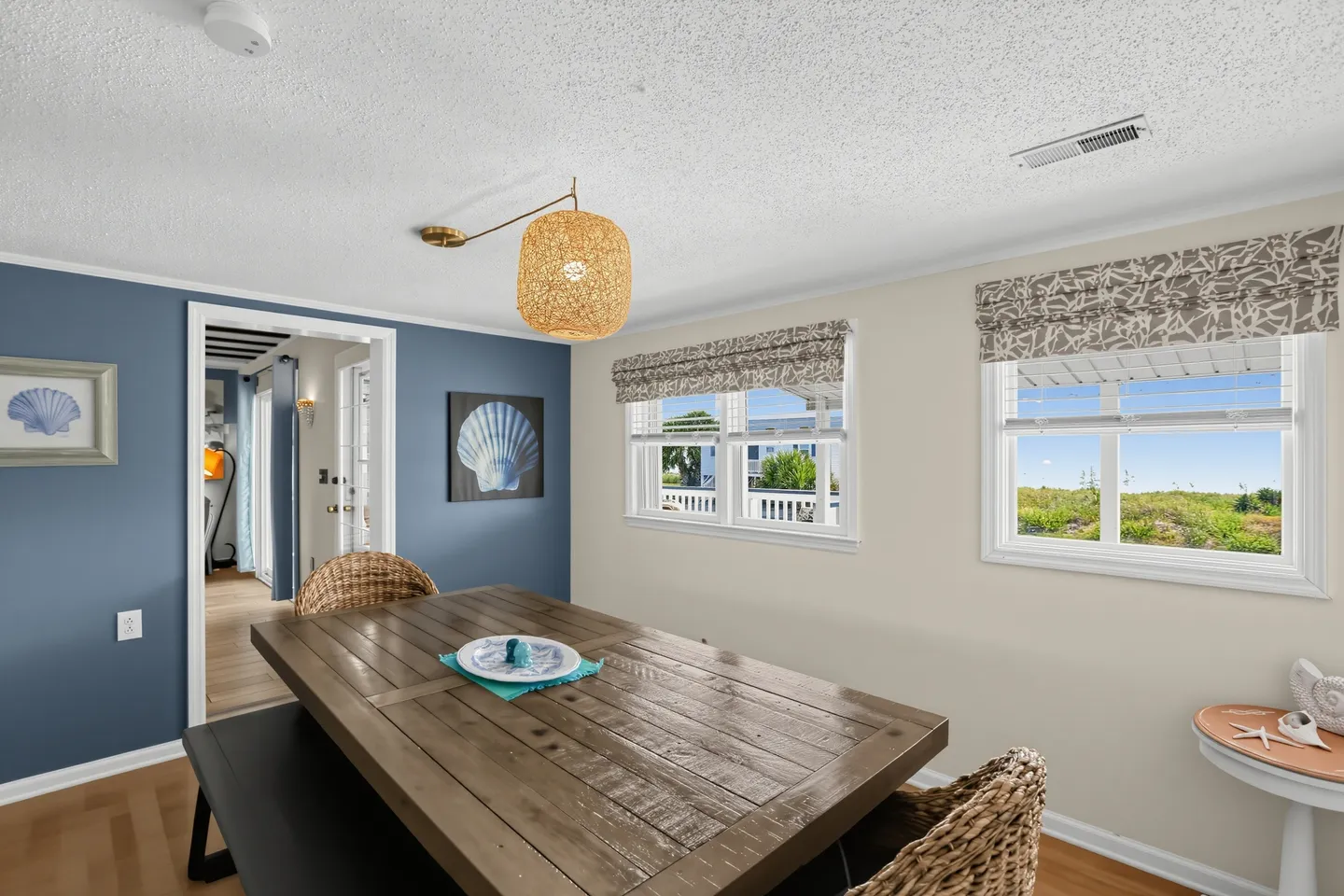 Dining area featuring a wooden table with wicker chairs, a bench, coastal artwork, and two windows with outdoor views.