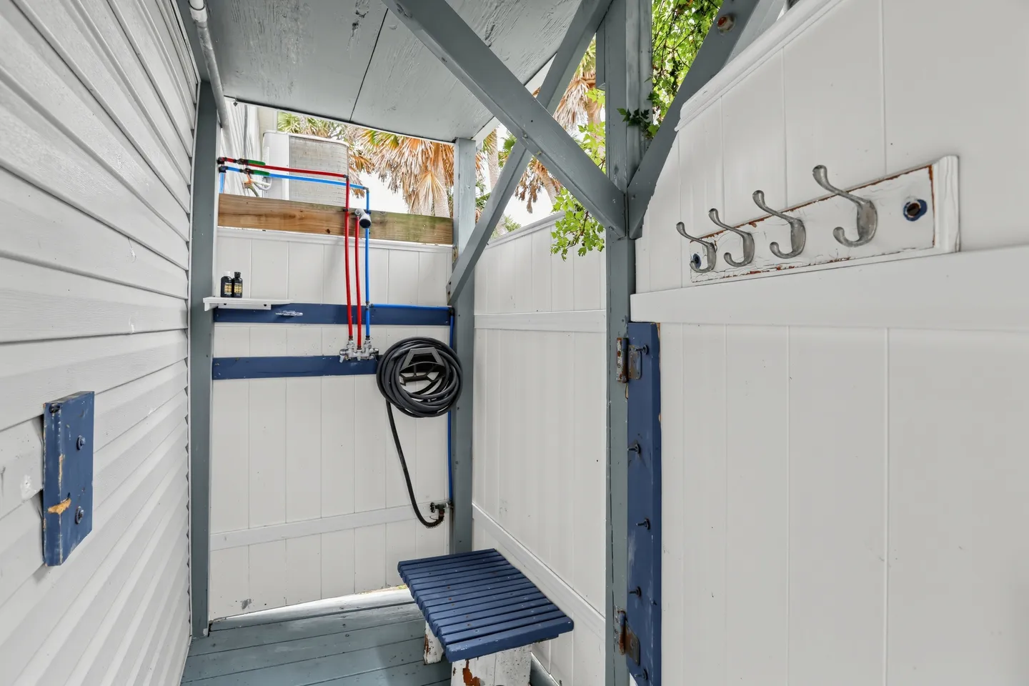 Enclosed outdoor shower area with white paneled walls, exposed plumbing, a hose, a small blue bench, and wall hooks.