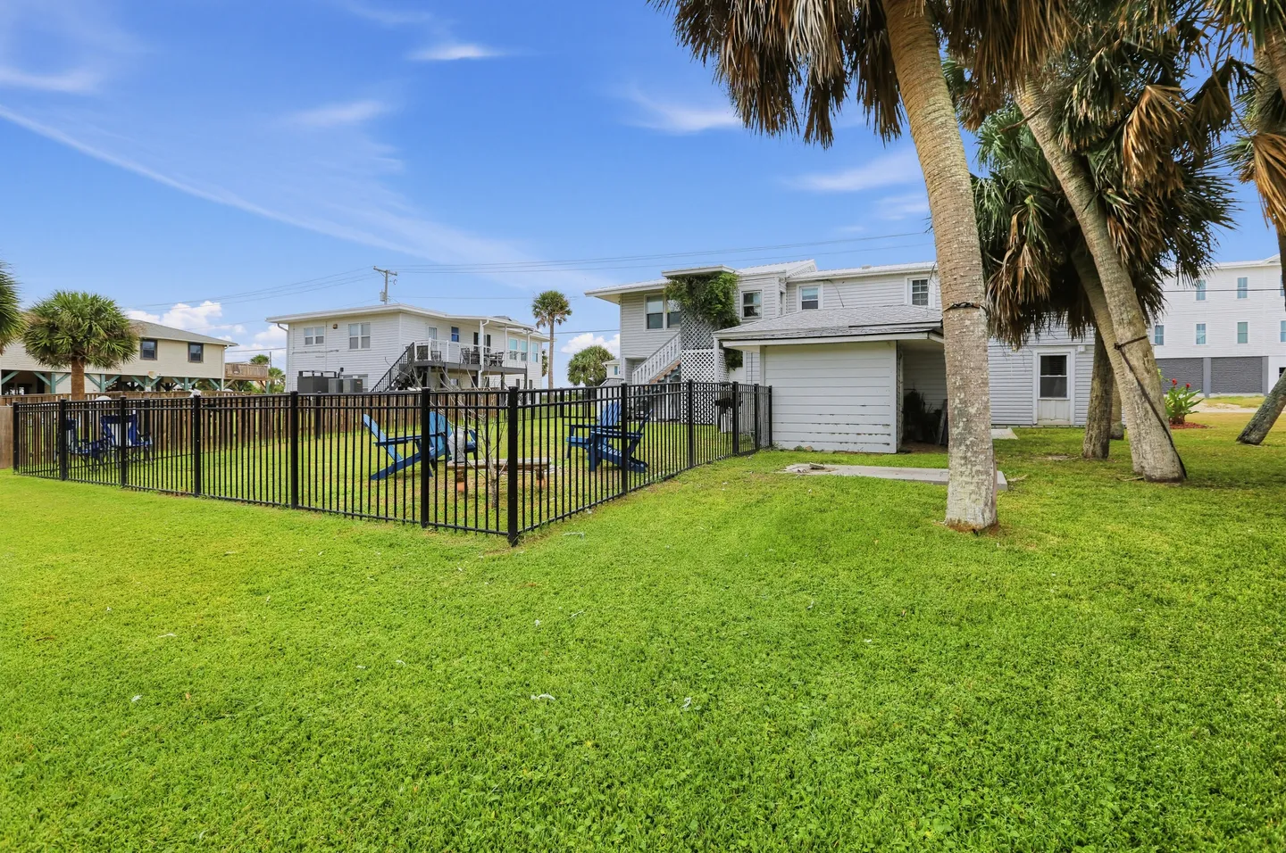 A grassy backyard featuring a black metal fenced-in patio area with blue Adirondack chairs and a fire pit under a blue sky.