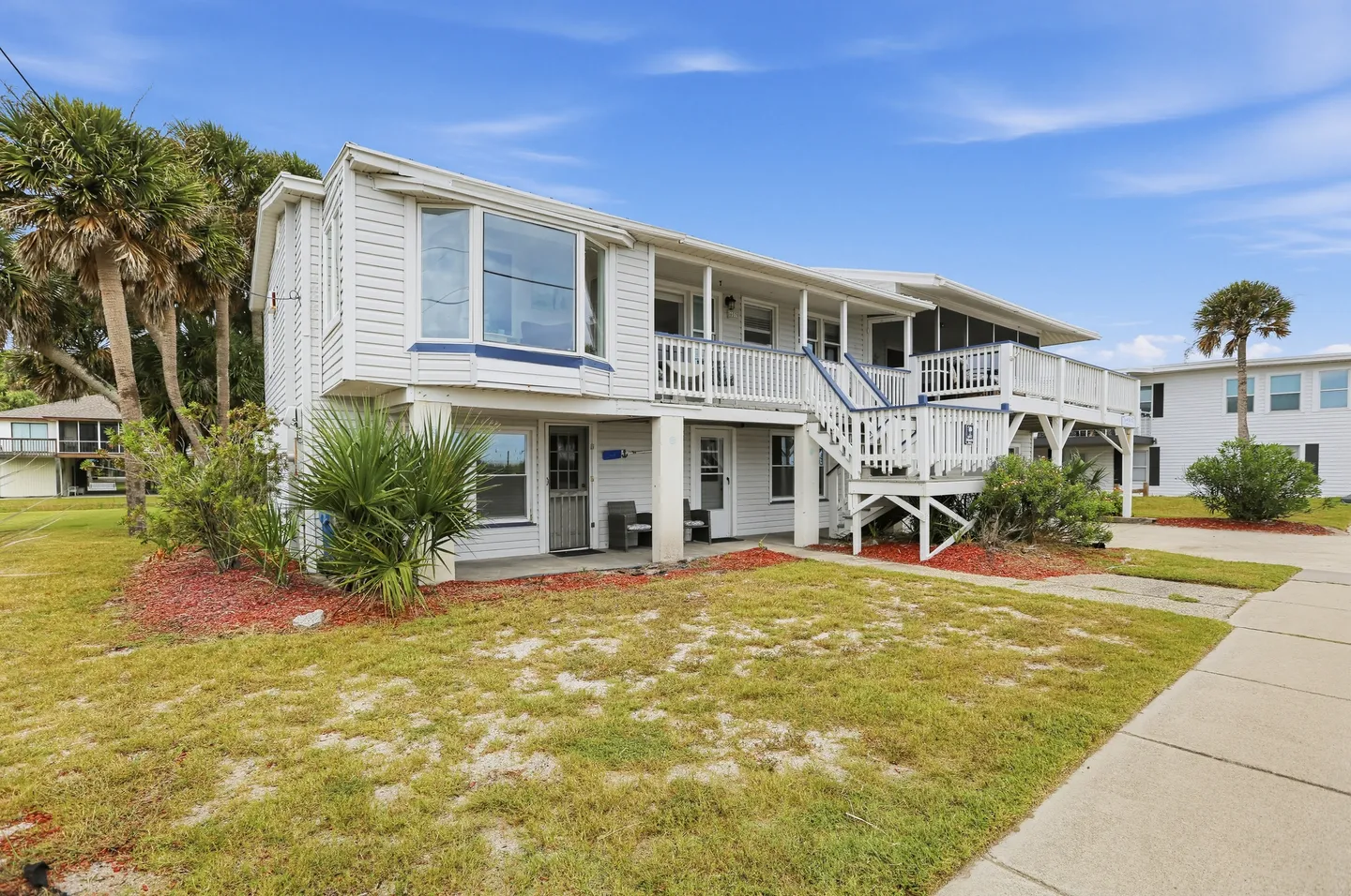 Exterior view of a two-story white house with upper-level balconies, exterior stairs, and surrounding lawn with palm trees under a blue sky.