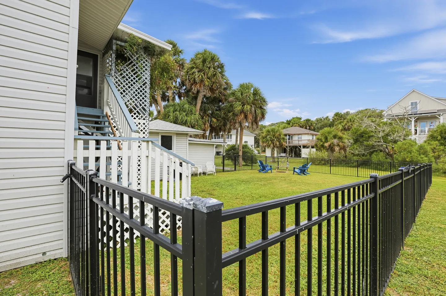 Fenced grassy backyard with blue chairs, palm trees, and an exterior staircase leading up the side of the house.