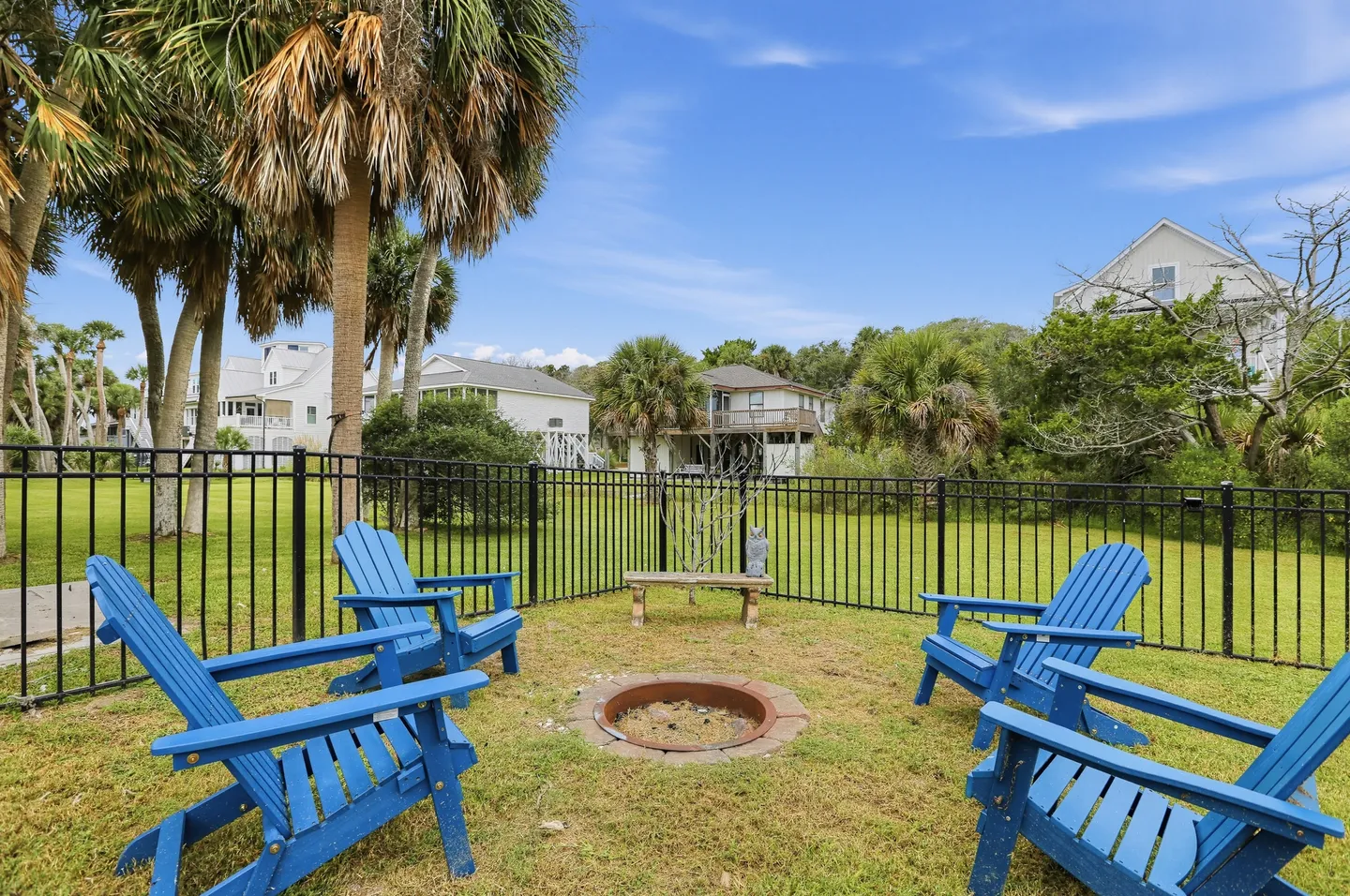 Fenced-in backyard seating area with blue Adirondack chairs surrounding an in-ground fire pit under palm trees.