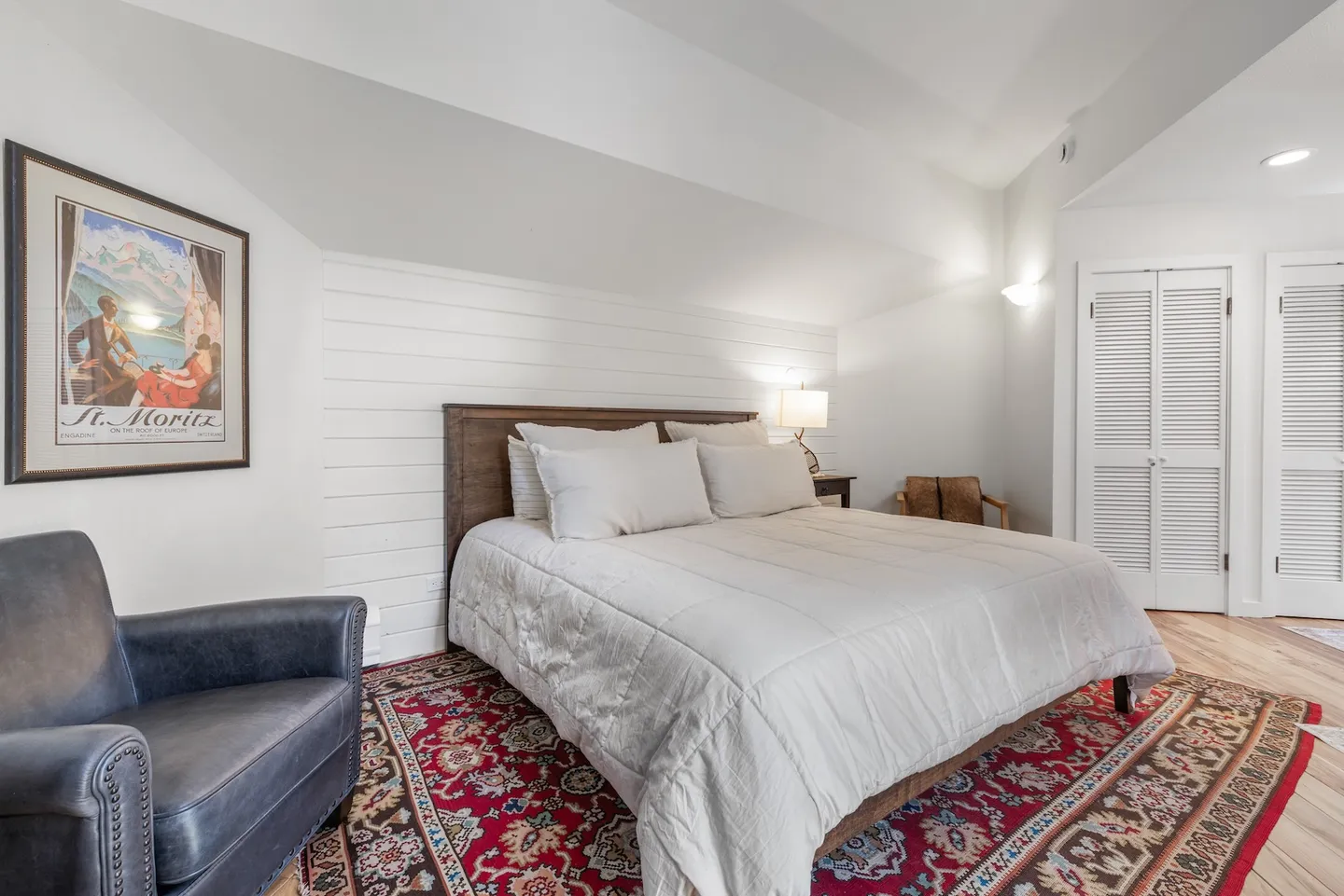 Bedroom with a wooden headboard, white bedding, an ornate red area rug, and an armchair under an angled ceiling.