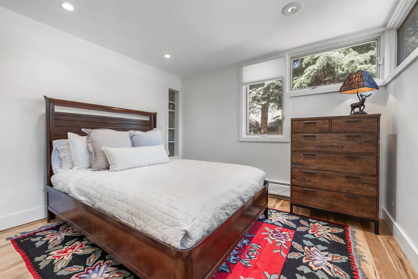 Bedroom featuring a wooden bed frame, patterned area rug, dresser with a deer lamp, and corner windows looking outside.