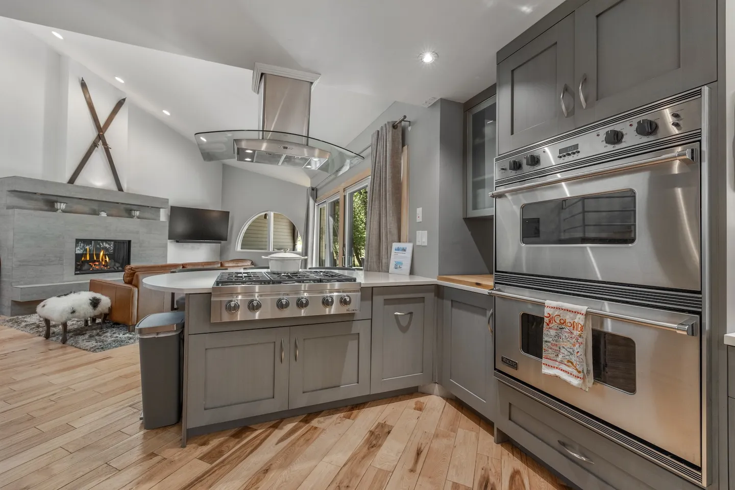 Kitchen island with stainless steel gas range and curved range hood, built-in double ovens, and view into the living room with fireplace.