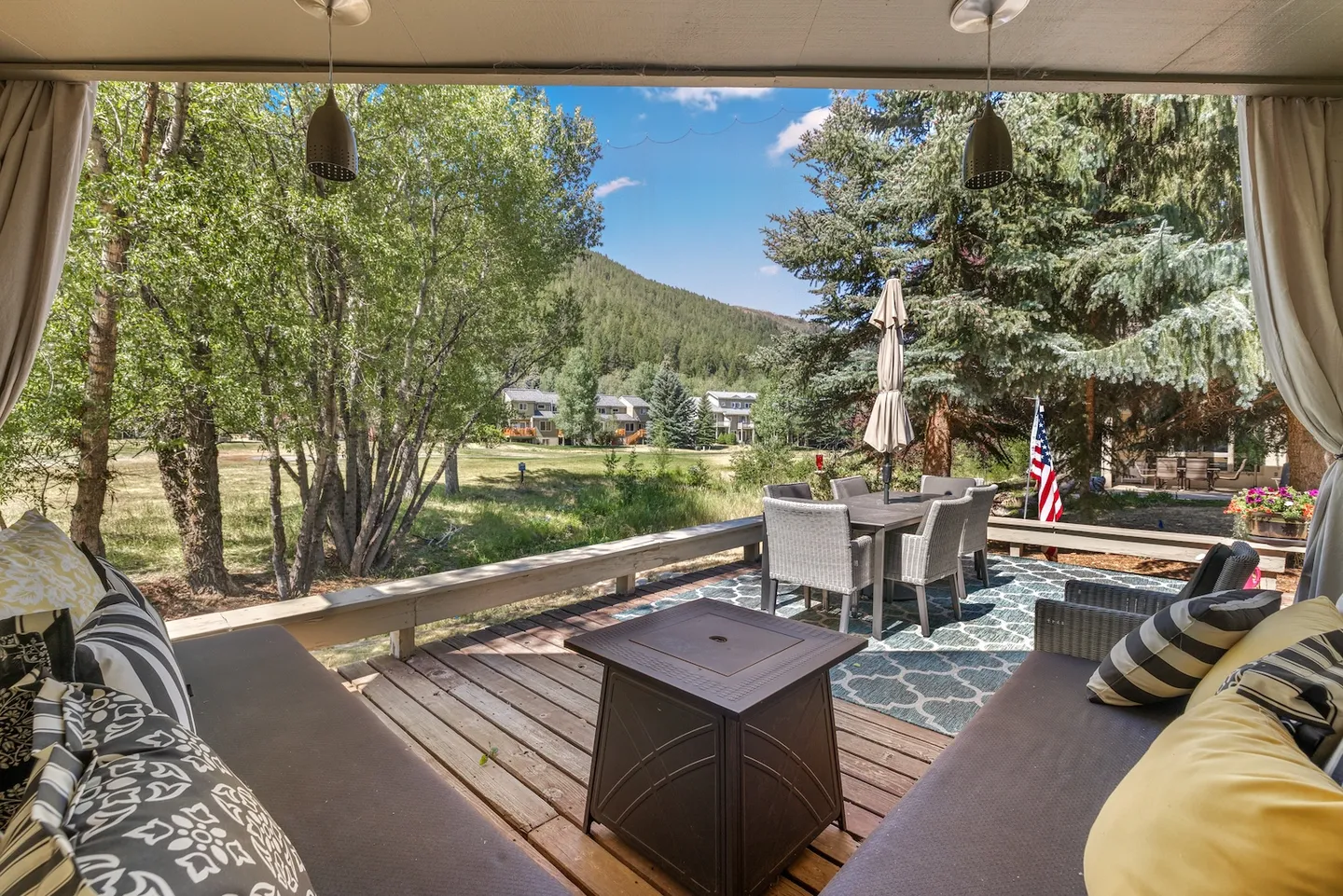 A covered deck with cushioned seating and a fire pit table, looking out onto an outdoor dining set, green trees, and mountain hills.