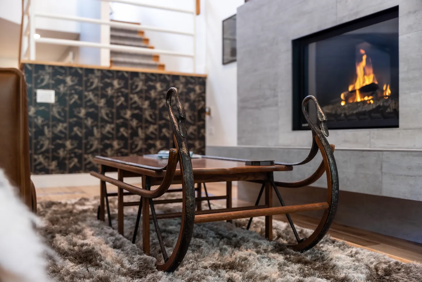 A wooden sled-style coffee table on a shag rug in front of a modern fireplace with a roaring fire.