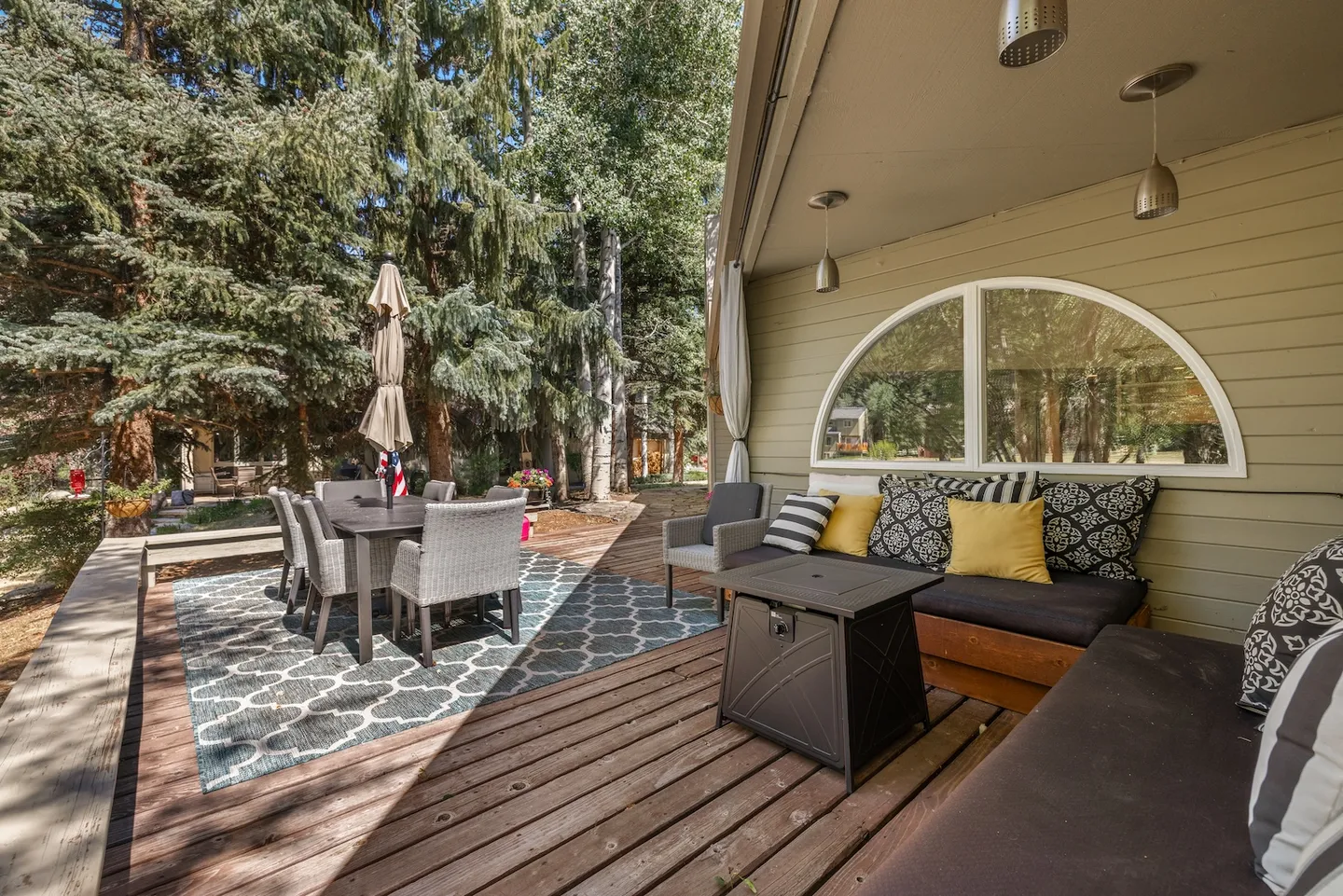 Outdoor wood deck featuring a covered lounge area with a sectional sofa and fire pit table, alongside a patio dining set on an outdoor rug surrounded by tall pine trees.