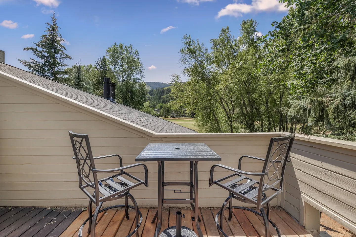 Outdoor balcony with two metal armchairs and a small square table overlooking surrounding trees and hills.