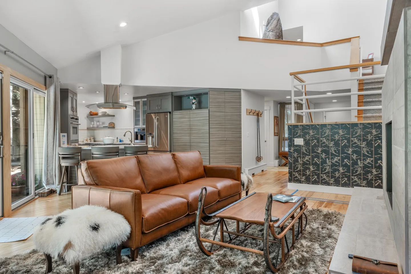 Open-concept living room featuring a brown leather sofa, a sled-style coffee table, and a kitchen with stainless steel appliances in the background.