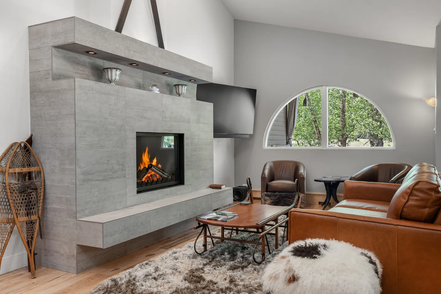 Living room with lit modern gray fireplace, leather seating, coffee table, and an arched window looking out to trees.