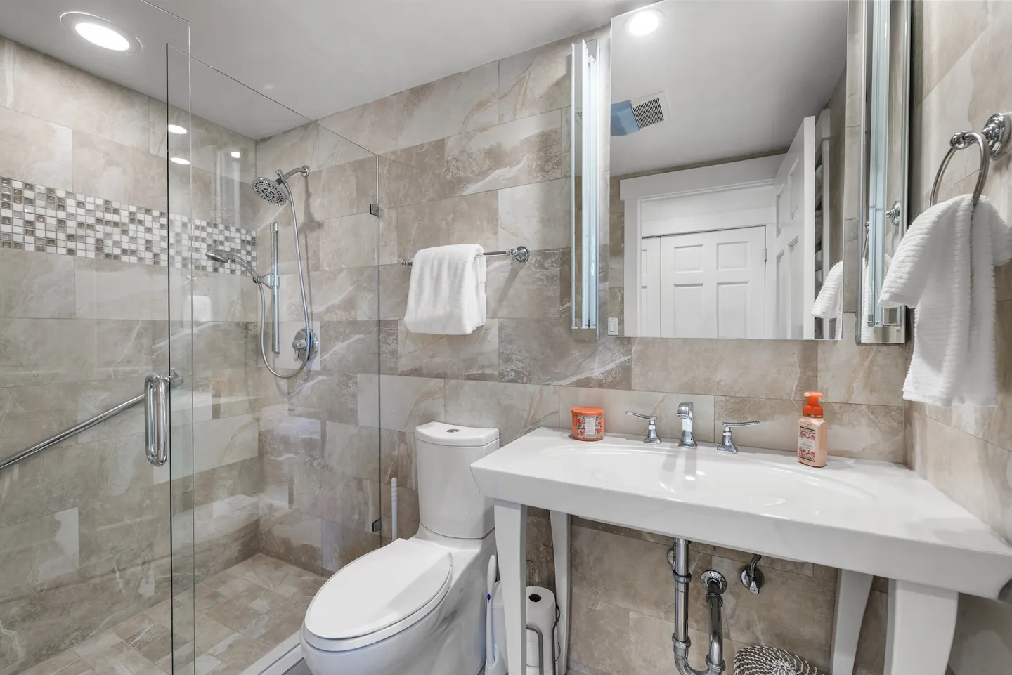 Bathroom with a glass-enclosed walk-in tile shower, toilet, and white console sink vanity with lighted mirror.