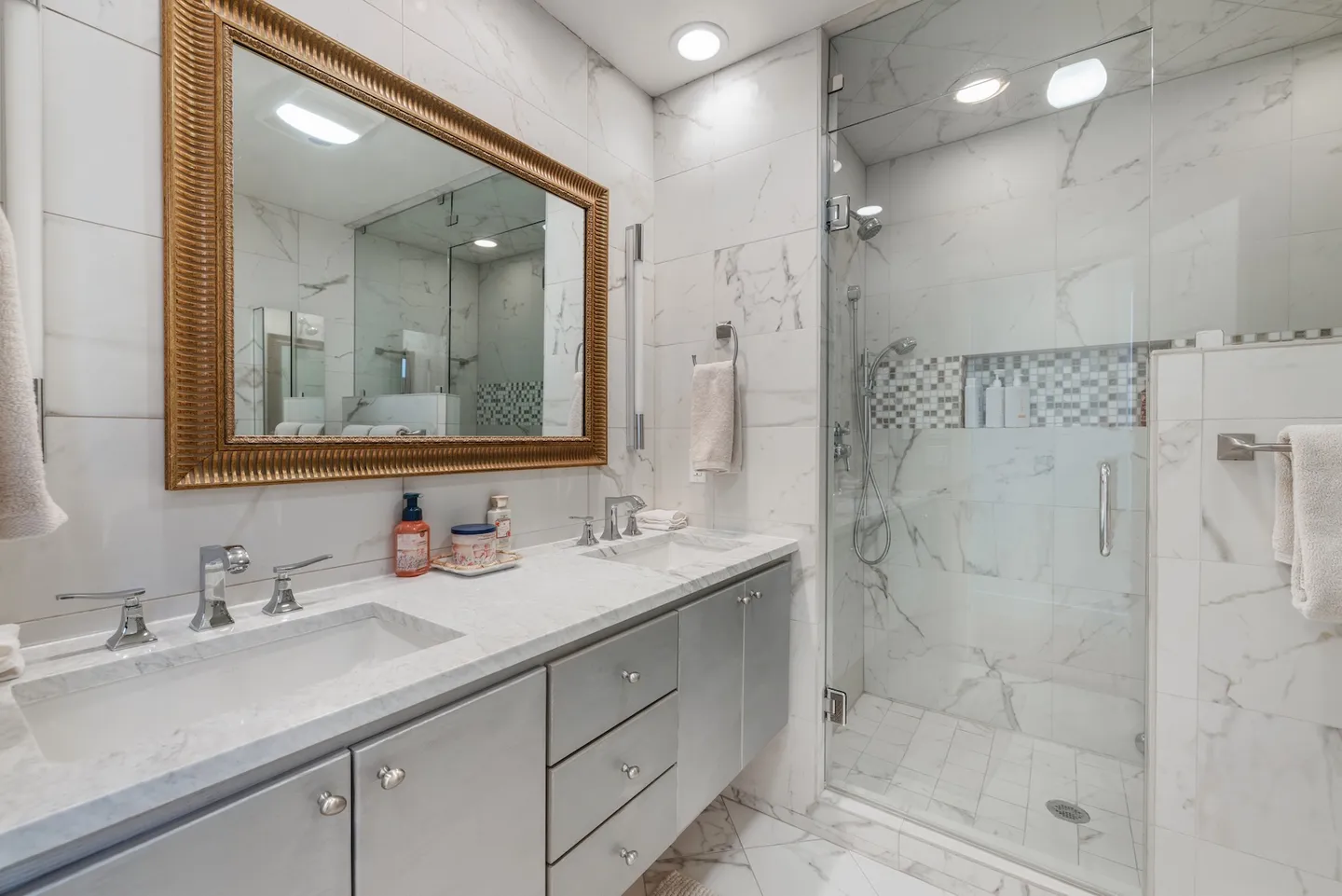 Bathroom with double-sink vanity, ornate gold-framed mirror, and glass-enclosed walk-in shower with marble-look tile.