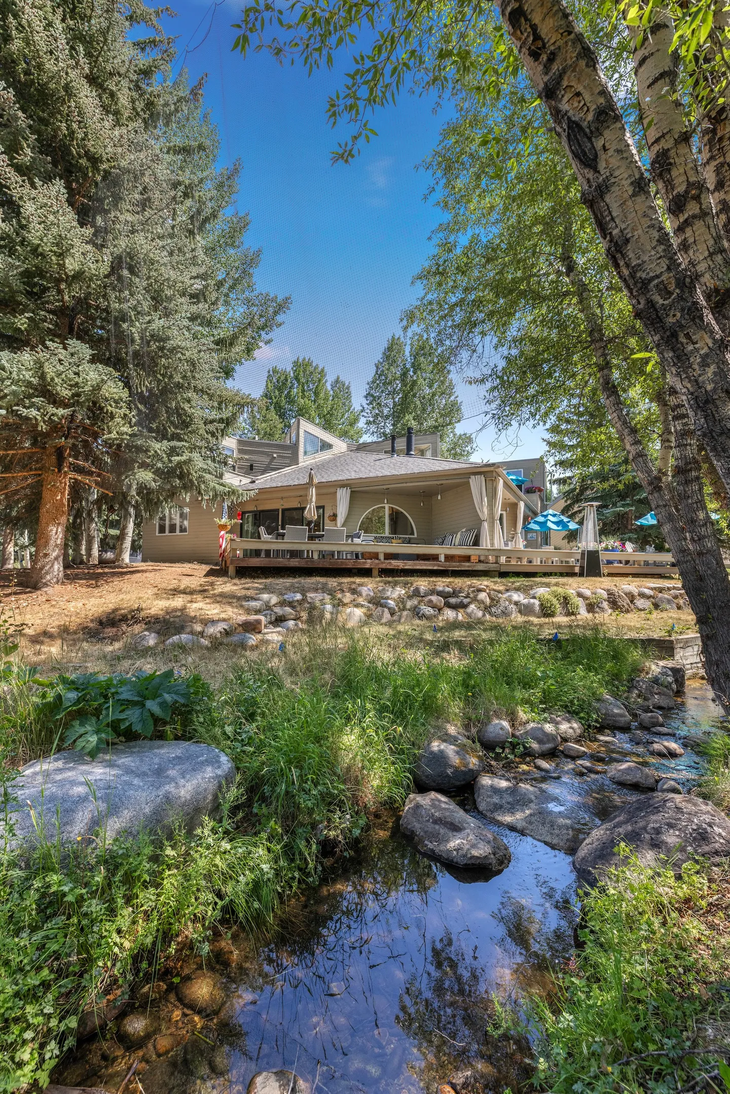 Rear exterior view of a multi-story house with a wooden back deck and gazebo structure, seen past a small rocky stream and surrounding trees.