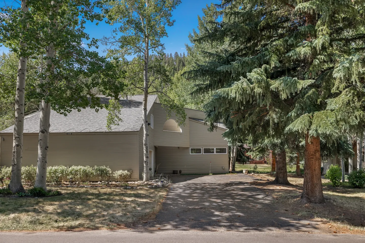 Exterior front view of a two-story home with light-colored siding, a paved driveway, and tall evergreen and aspen trees under a blue sky.