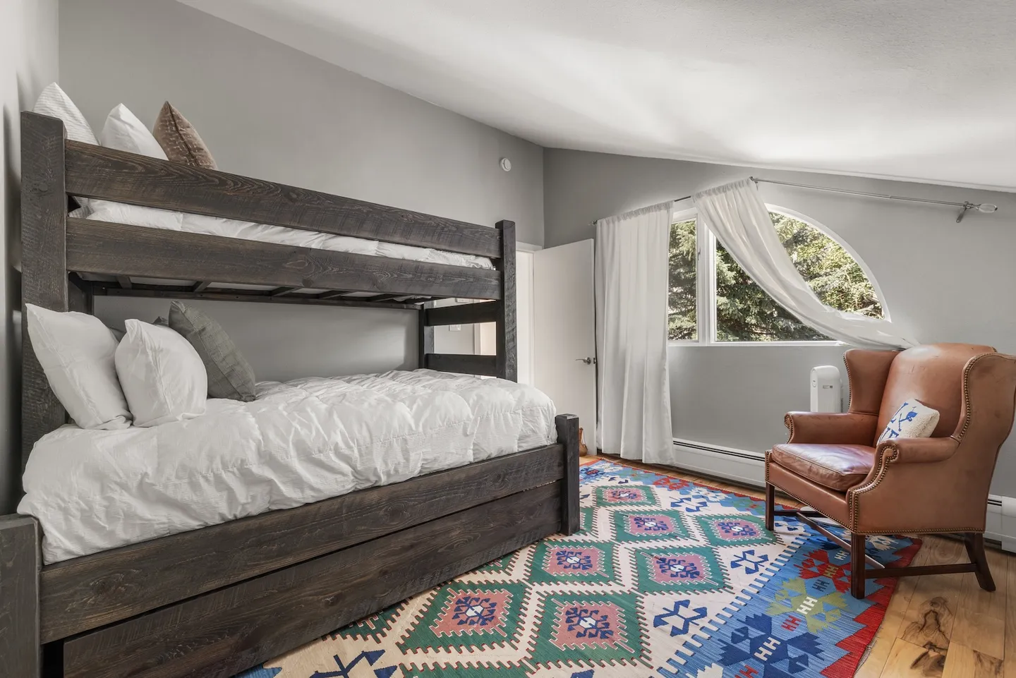 Bedroom with a rustic dark wood bunk bed, a colorful patterned rug, and a leather armchair beneath an arched window.