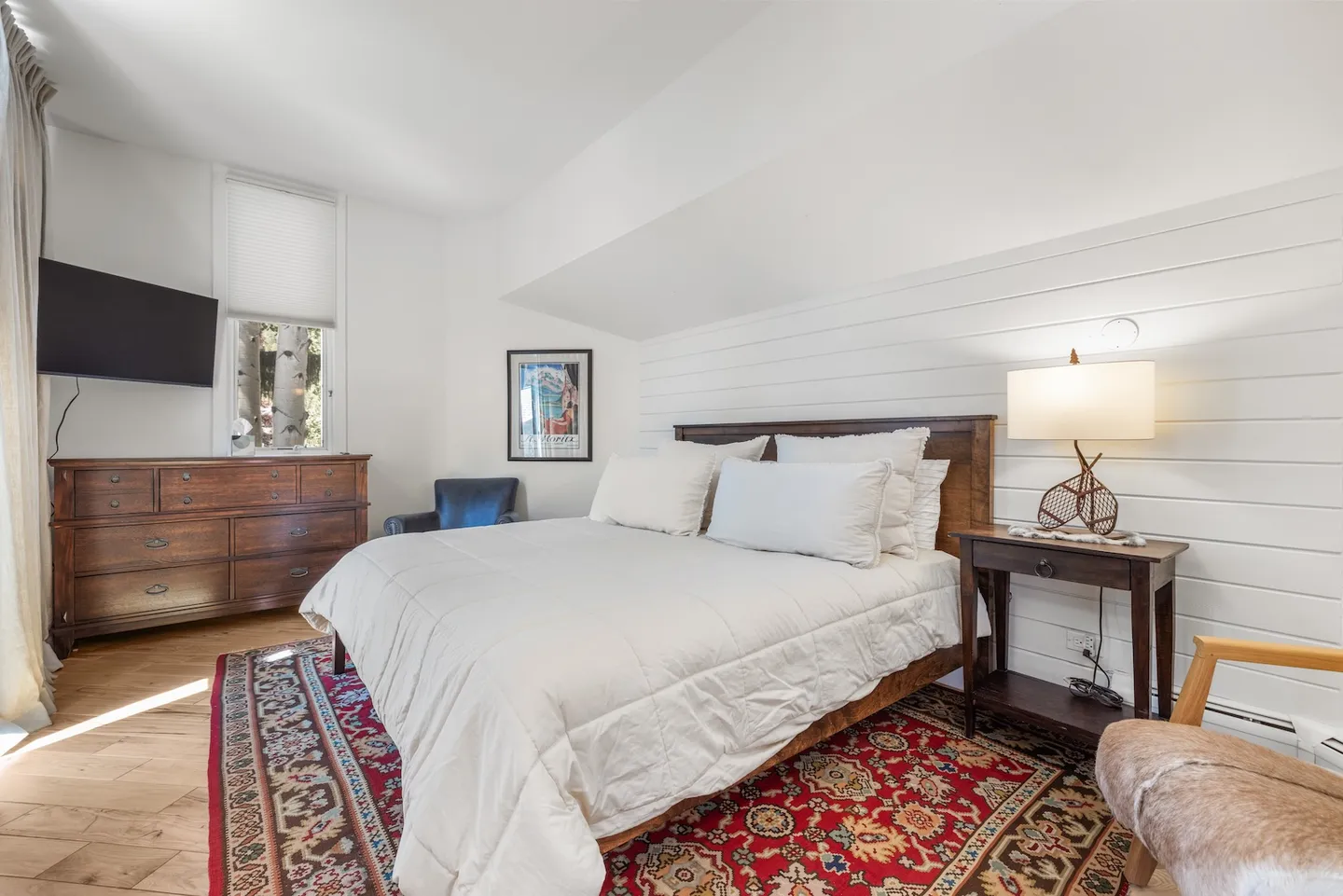 Bedroom with a wooden bed, white bedding, oriental-style area rug, nightstand with lamp, and a dresser below a wall-mounted TV.