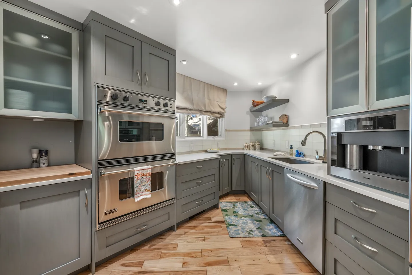 Kitchen with gray cabinetry, light countertops, wood flooring, and built-in stainless steel appliances including double ovens and a built-in coffee maker.