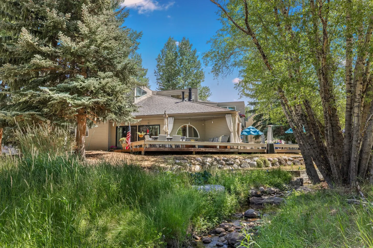 Exterior view of a home with a wooden deck bordered by trees, a rocky bank, and a small stream in the foreground.