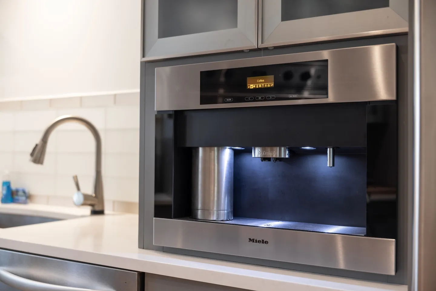 Close-up of a stainless steel built-in Miele coffee machine integrated into kitchen cabinetry near a sink.