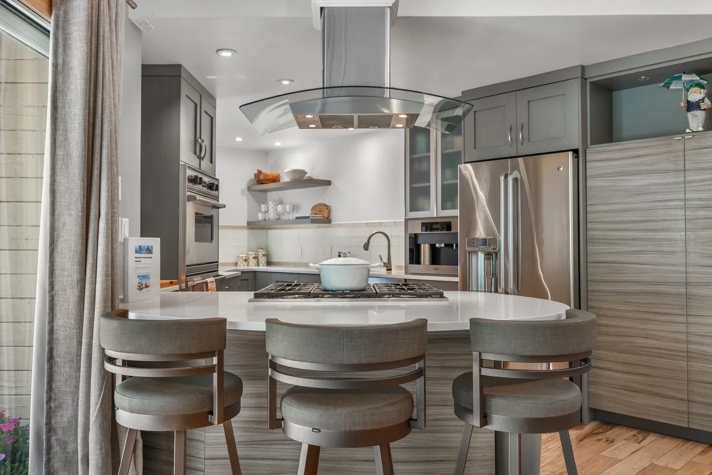 A modern kitchen island with a curved glass vent hood, built-in cooktop, and three padded barstools in front of gray cabinetry and stainless steel appliances.