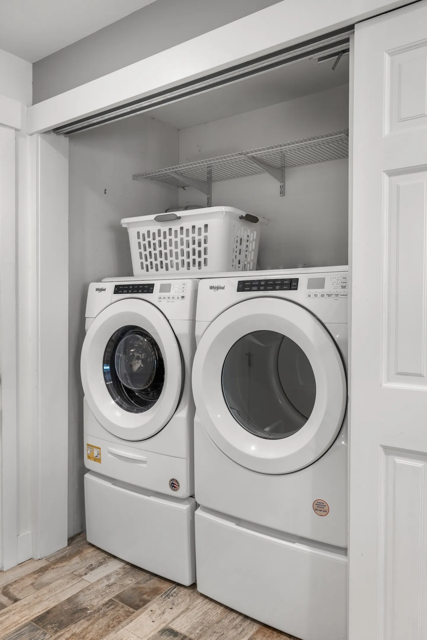 A laundry closet featuring a side-by-side front-loading Whirlpool washer and dryer with pedestal drawers, a wire shelf, and a white laundry basket.