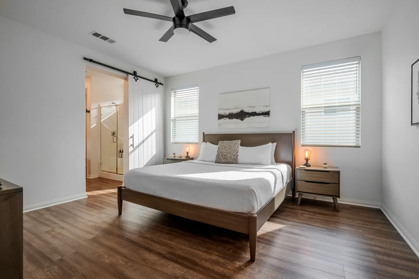 Bedroom featuring a wooden bed frame, two nightstands with small lamps, wood-look flooring, a ceiling fan, and an open sliding barn door to the bathroom.