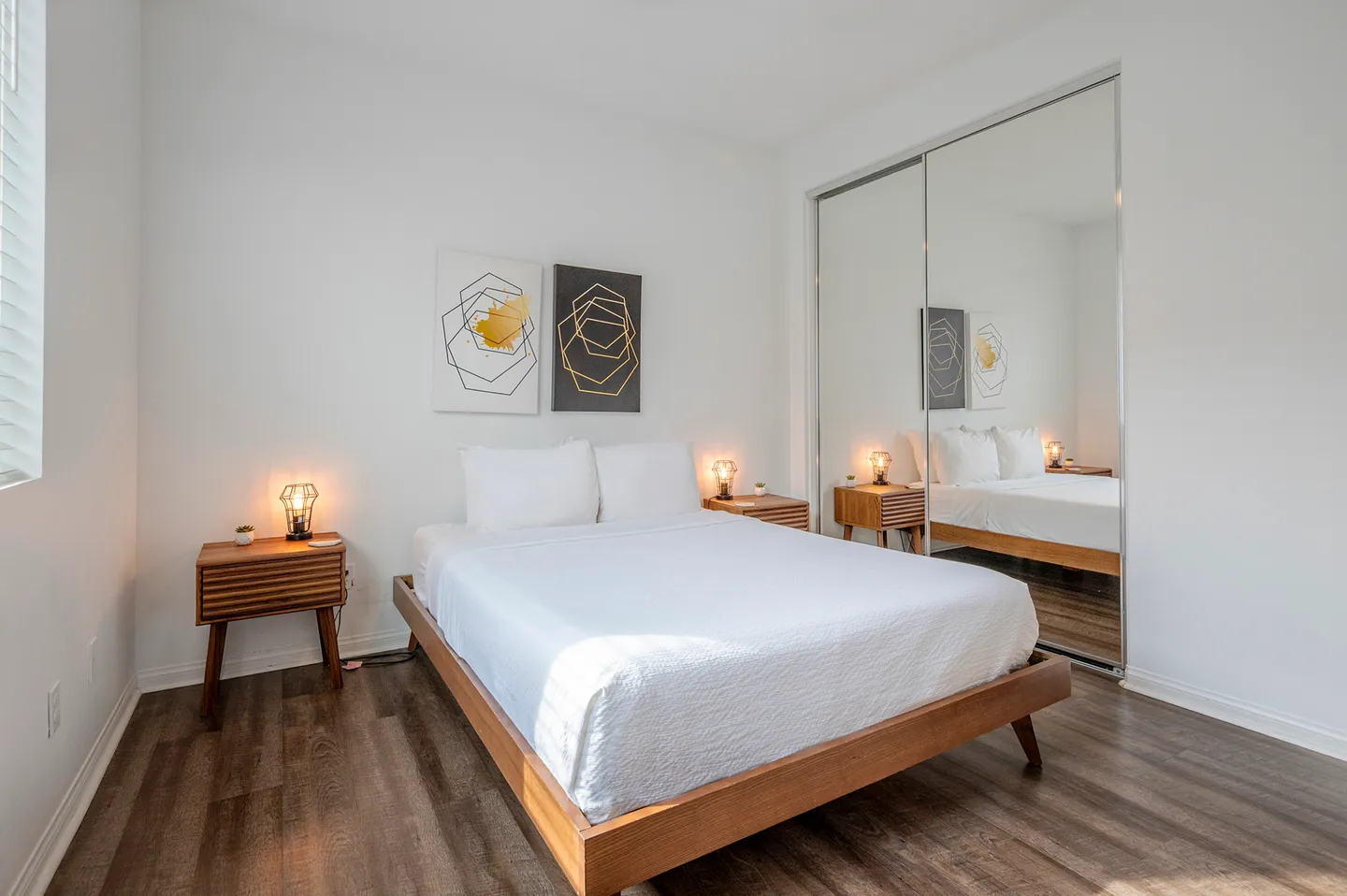 Bedroom with a platform bed dressed in white linens, wood nightstands with small lamps, geometric wall art, and mirrored closet doors.