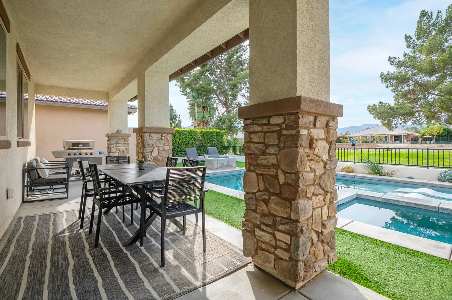 Covered patio featuring an outdoor dining table and seating, supported by stone-accented pillars, overlooking a swimming pool and yard.