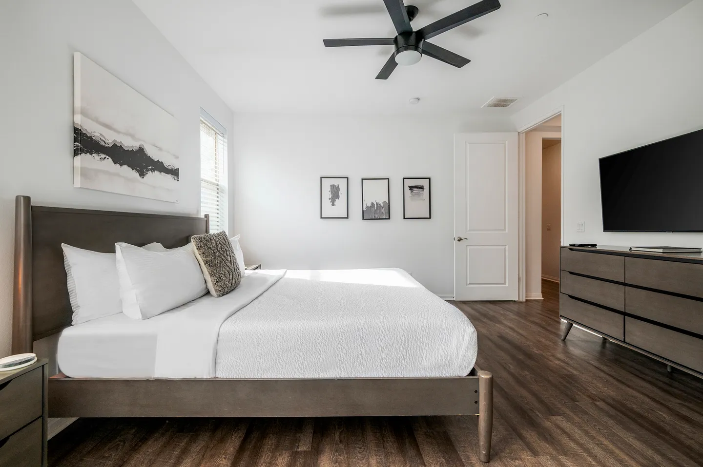 Bedroom with a wooden bed frame, white bedding, ceiling fan, wall-mounted television, and a dresser on dark wood-look floors.