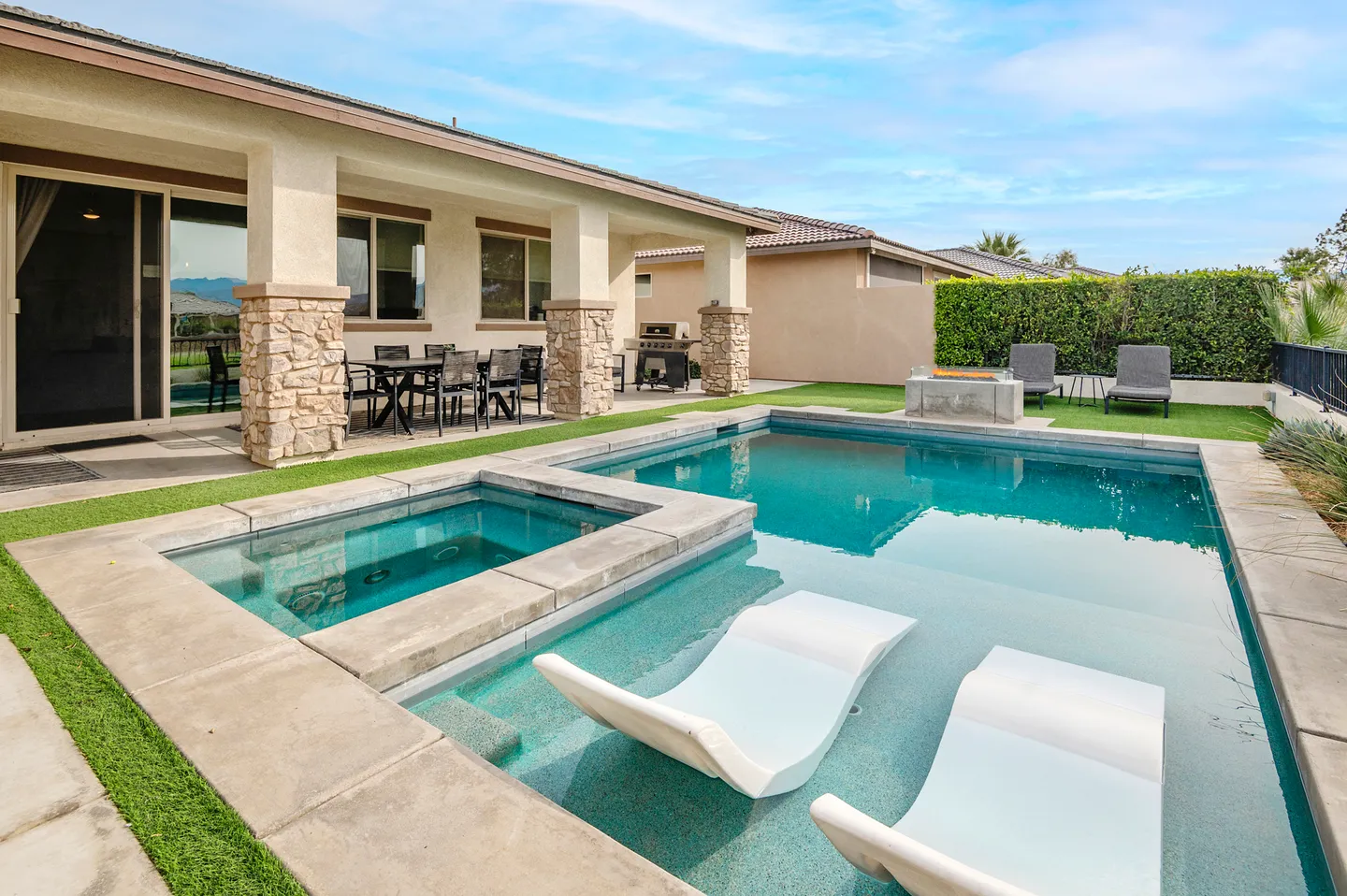 Outdoor swimming pool with integrated hot tub, in-pool lounge chairs, covered patio, and fire pit in a backyard.