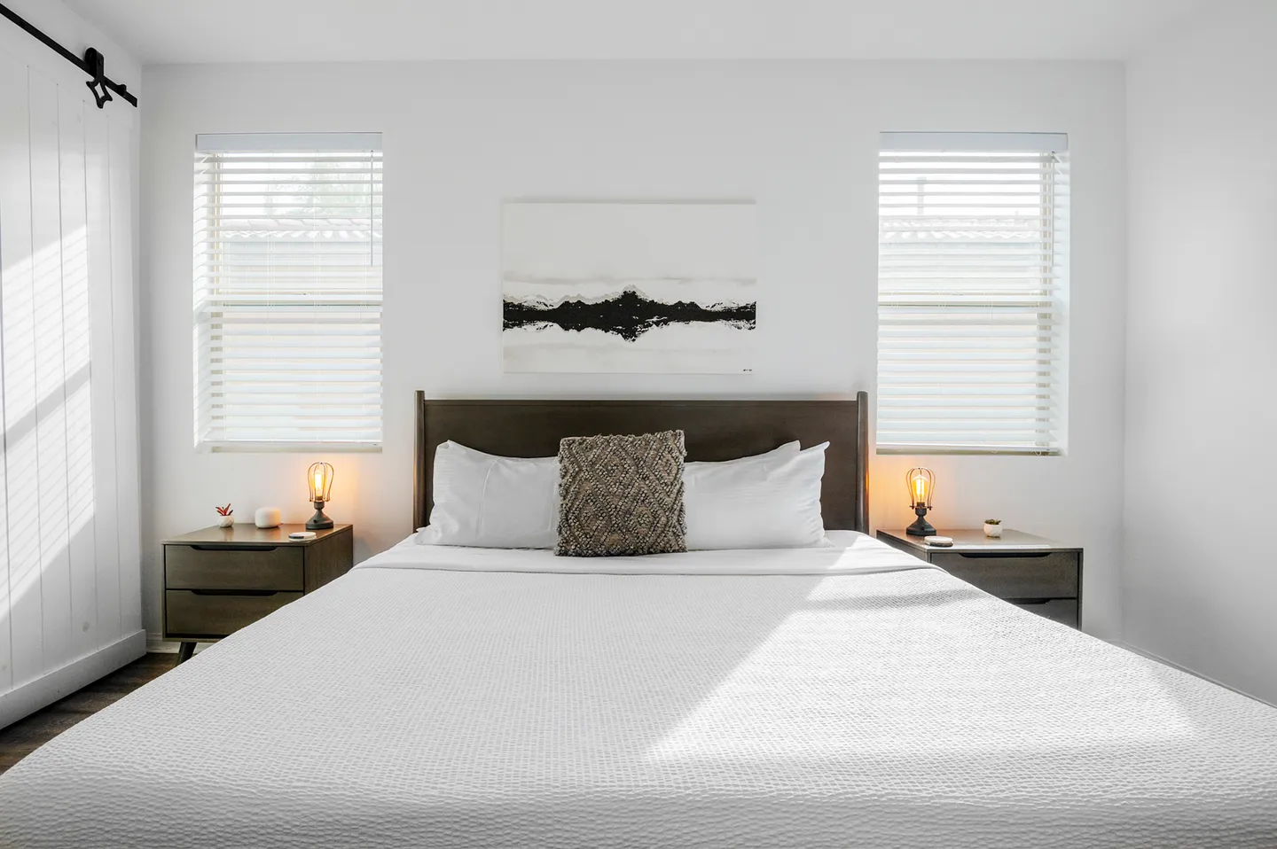 Bedroom with a bed dressed in white linens flanked by two nightstands and windows with blinds.