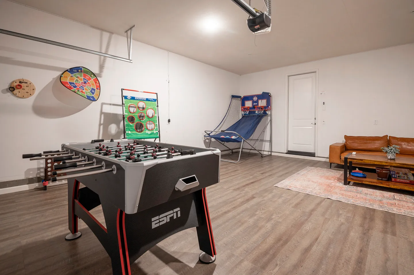 Game room setup inside a converted garage space featuring a foosball table, an arcade basketball game, and a leather sofa.