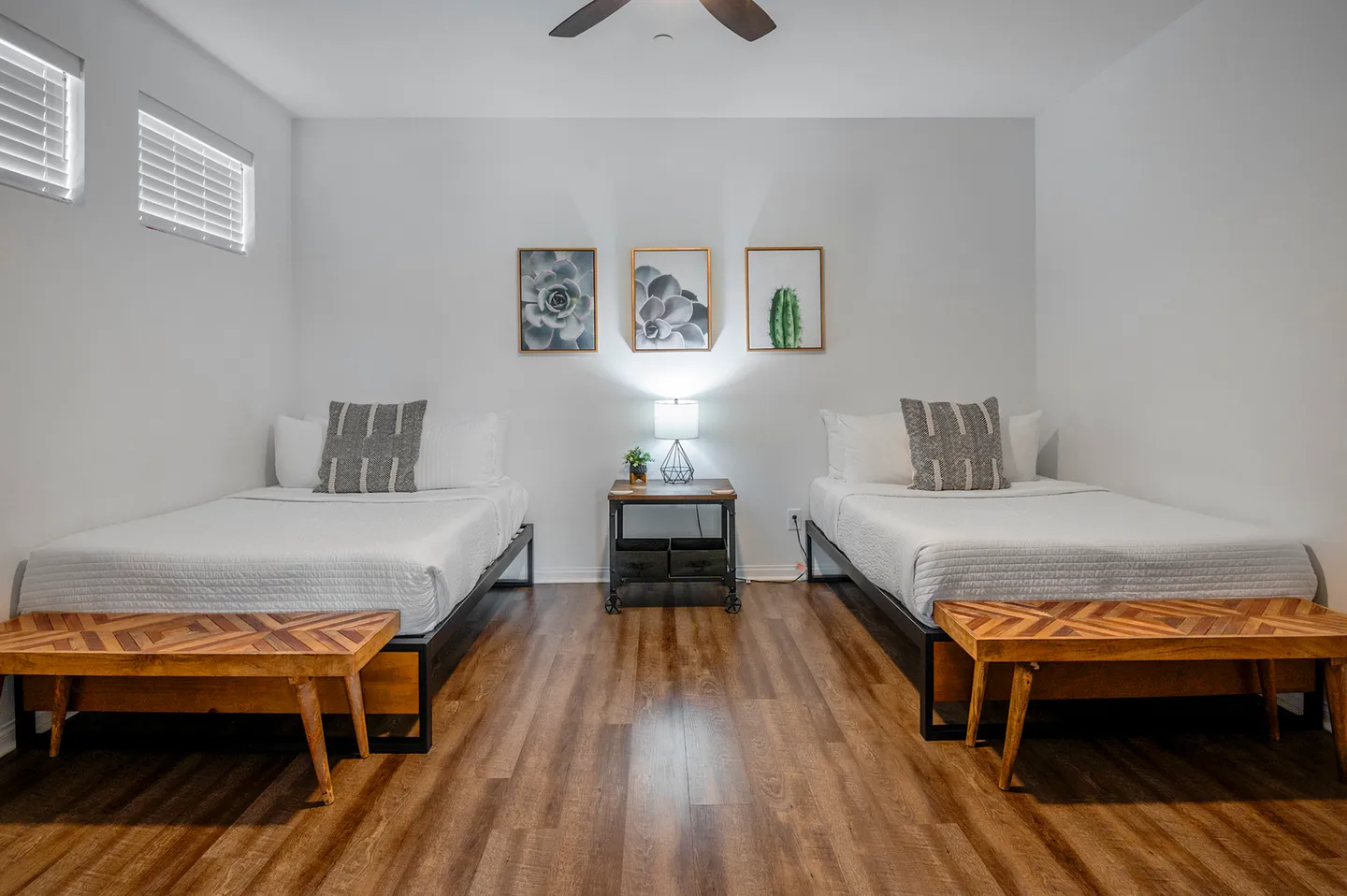 Bedroom featuring two beds with patterned accent pillows and wooden benches at the foot of each, separated by a nightstand with a lamp.