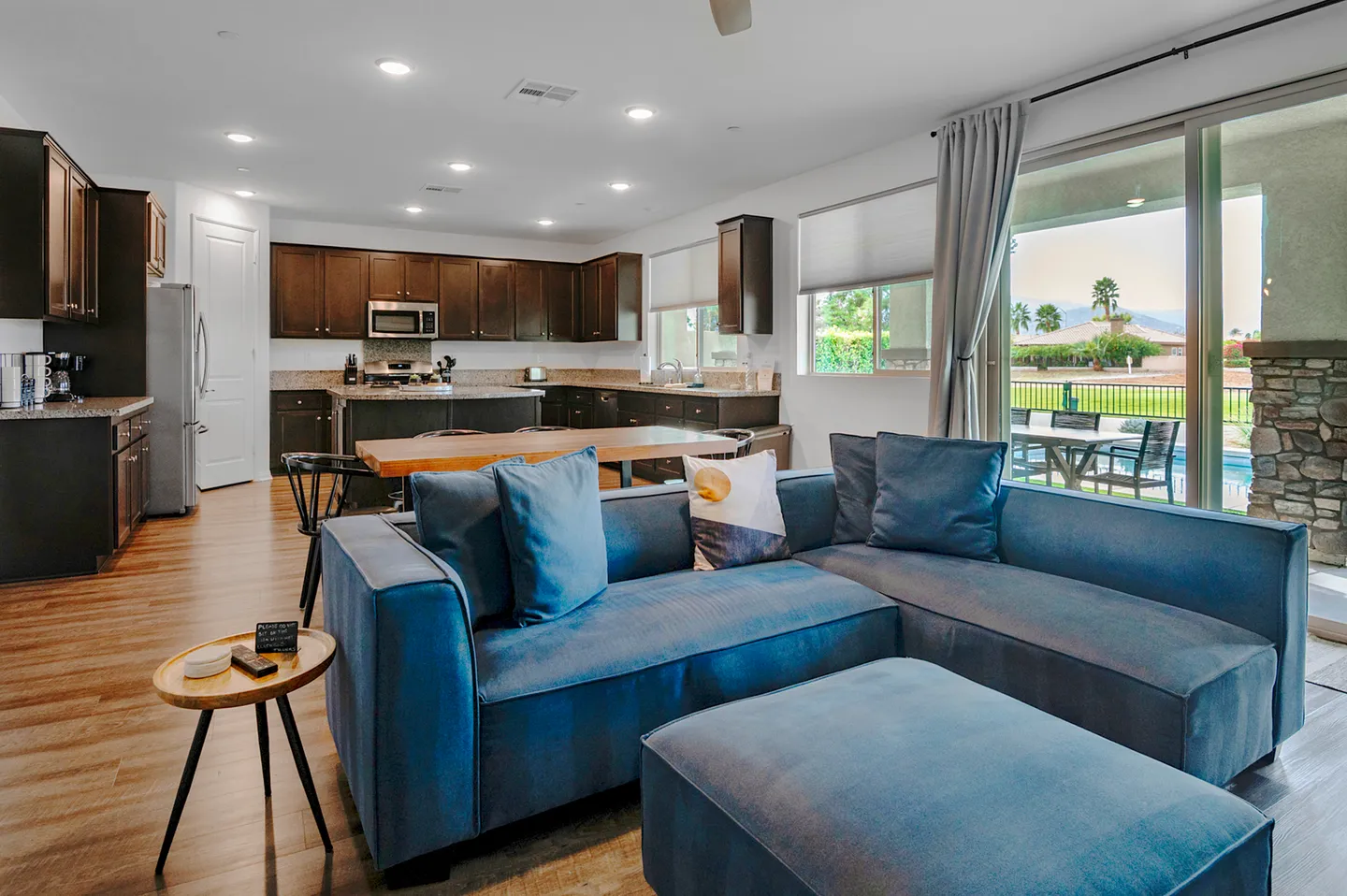 Open-concept living room with a blue sectional sofa and ottoman adjacent to a kitchen with dark cabinetry and sliding glass doors leading outside.