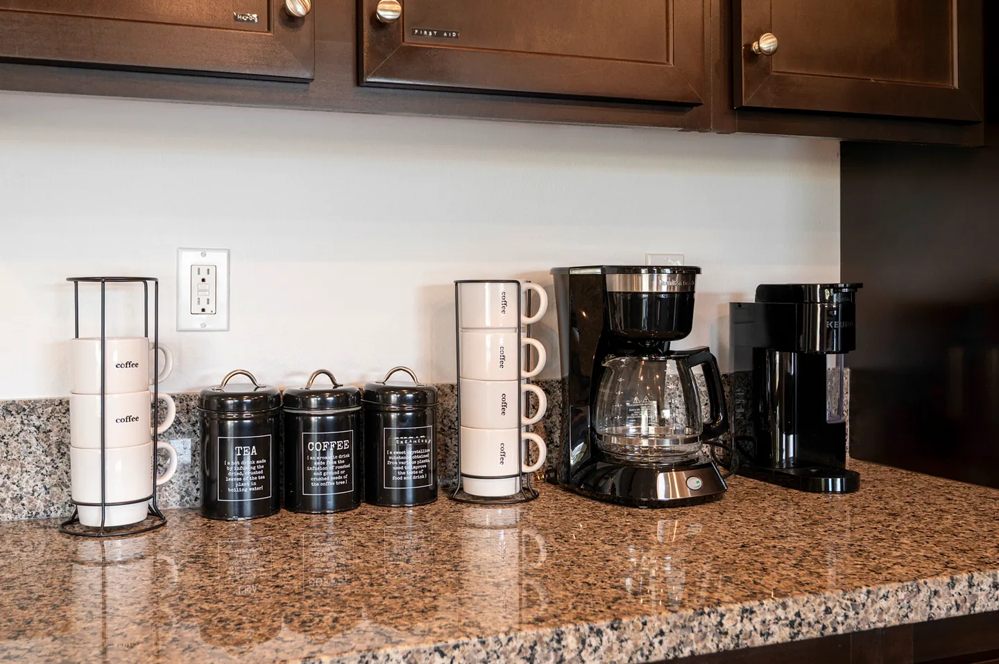 Close-up of a kitchen countertop with two coffee makers, black canisters, and stacked coffee mugs beneath dark wood cabinets.