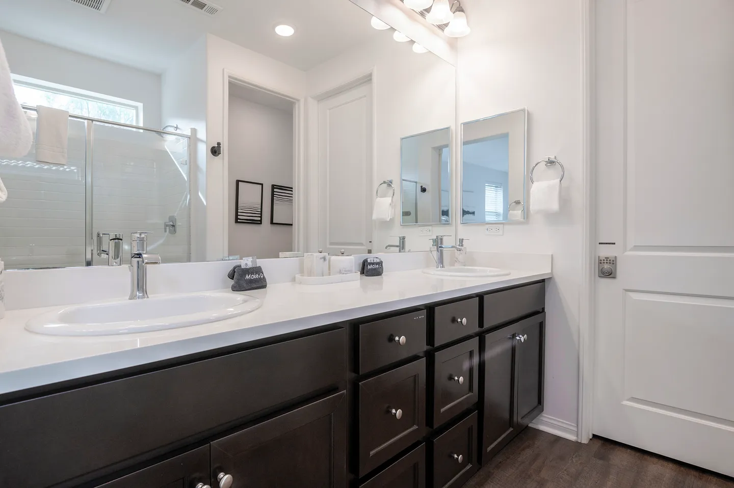 Bathroom with a double-sink vanity, dark cabinets, a large mirror, and a walk-in shower with glass doors visible in the reflection.
