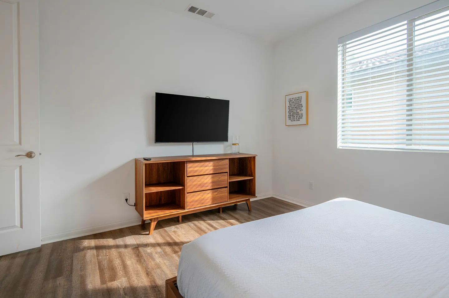 Bedroom with wood-look flooring, a wall-mounted flat-screen TV above a wooden console table, and a bed corner in the foreground.