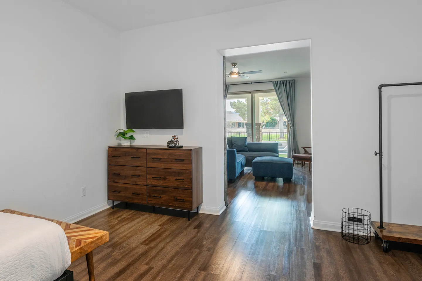 Bedroom with wood-style flooring, a wooden dresser beneath a wall-mounted TV, and a doorway looking into an adjoining living area.