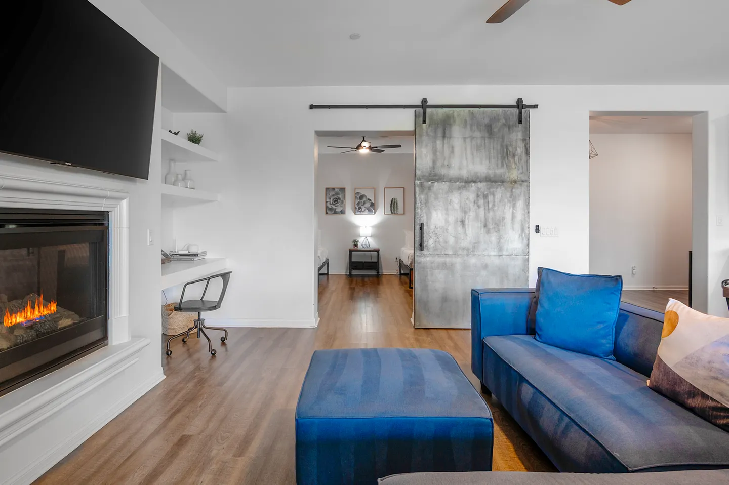 Living room featuring a lighted fireplace beneath a TV, a blue sofa with ottoman, and a sliding metal barn door leading to an adjacent bedroom.