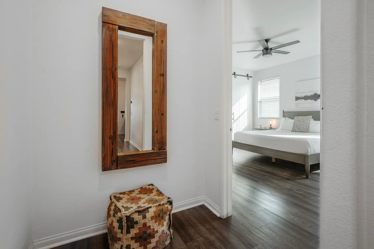 Hallway alcove with a rustic wooden mirror and patterned ottoman, looking into a sunlit bedroom.